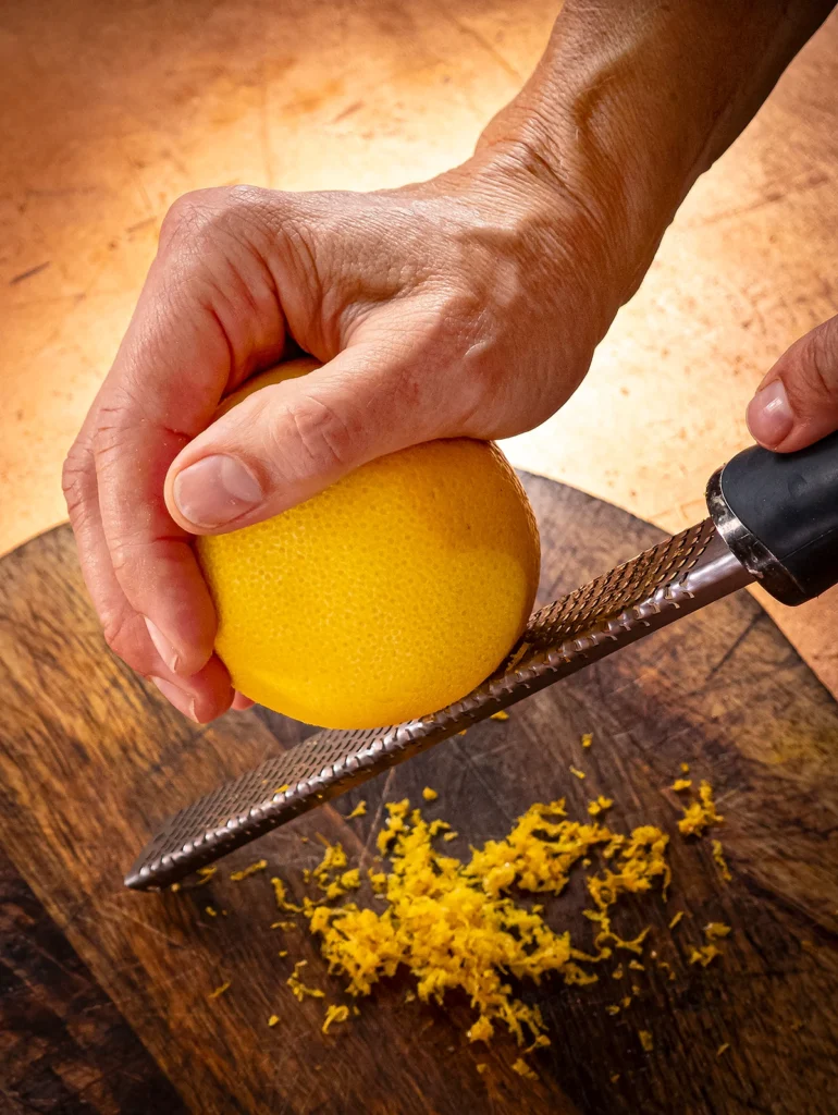 A wooden cutting board stands on a bronze-colored base. One hand holds an orange and rubs it over a zester. A heap of orange peel lies on the wooden board.