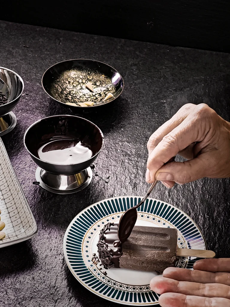 A hand holds a spoon with melted chocolate to place a blob to the eye area on the popsicle that lays on a plate. Behind it are three bowls of melted chocolate, chocolate sprinkles, and almond pieces prepared for the teeth decoration. Everything stands on a black slate background.