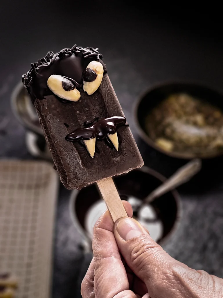 One hand holds a Halloween popsicle with sesame seeds. In the background, slightly blurred, you can see the bowls with the ingredients for the decoration.