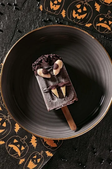 A Halloween sesame popsicle lies on a black plate with a gold rim. On the left-hand side and behind are napkins patterned with Halloween pumpkins. Beluga lentils are scattered on the black slate background.