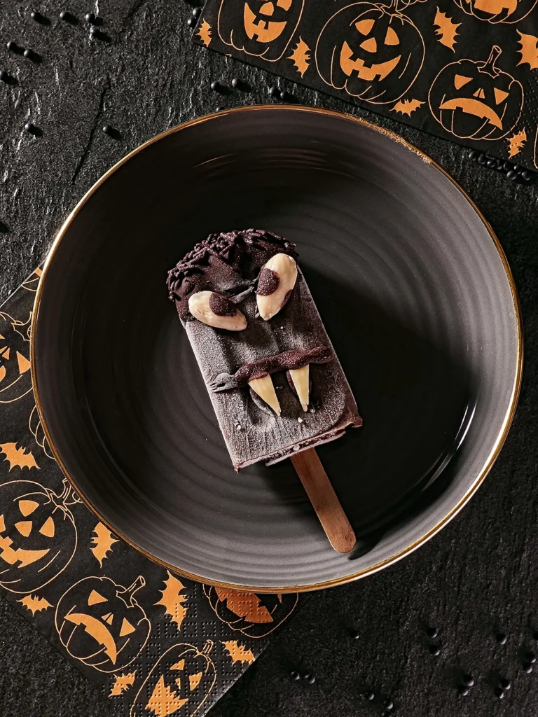 A Halloween sesame popsicle lies on a black plate with a gold rim. On the left-hand side and behind are napkins patterned with Halloween pumpkins. Beluga lentils are scattered on the black slate background.