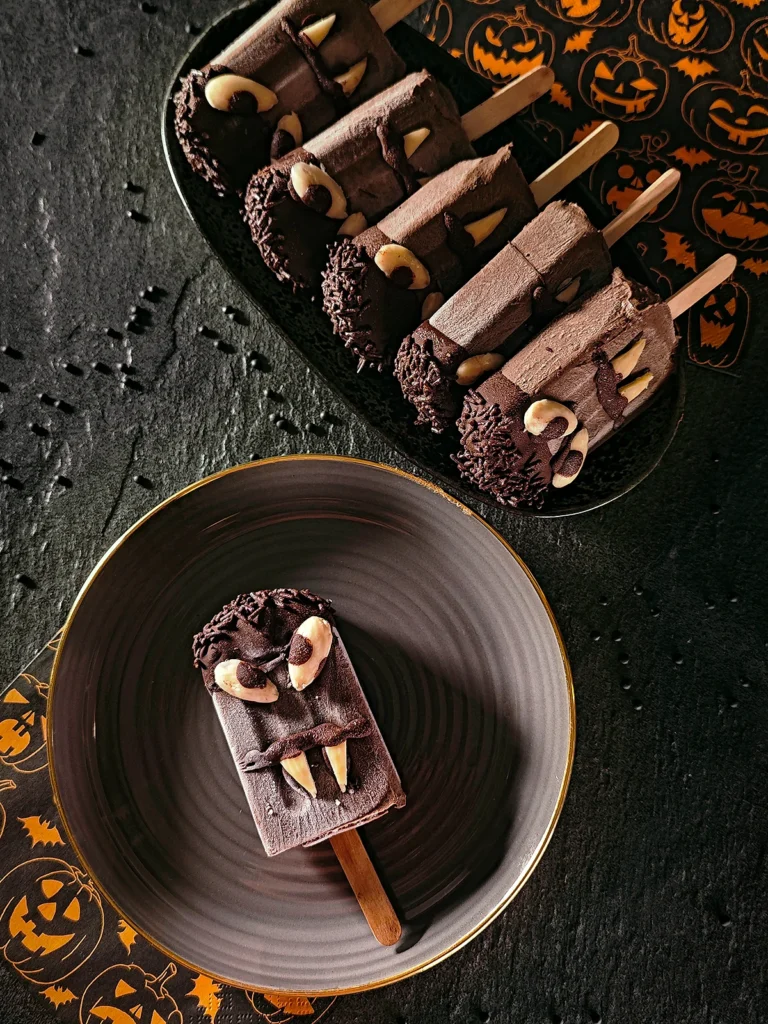 A Halloween sesame popsicle lies on a black plate with a gold rim. On the left-hand side is a napkin patterned with Halloween pumpkins. Behind it is a black oblong bowl with five more Halloween sesame popsicles. A napkin with a Halloween pumpkin pattern can also be seen behind it. Beluga lentils are scattered on the black slate background.