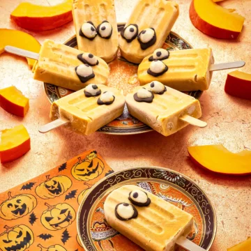 On a bronze-colored background, a small patterned plate with a Halloween popsicle with pumpkin lies in the foreground. Next to it is a napkin printed with pumpkin motifs. Behind it is a larger plate with six more Halloween popsicles. Surrounded by slices of sliced Hokkaido pumpkin.