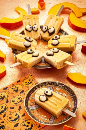 On a bronze-colored background, a small patterned plate with a Halloween popsicle with pumpkin lies in the foreground. Next to it is a napkin printed with pumpkin motifs. Behind it is a larger plate with six more Halloween popsicles. Surrounded by slices of sliced Hokkaido pumpkin.