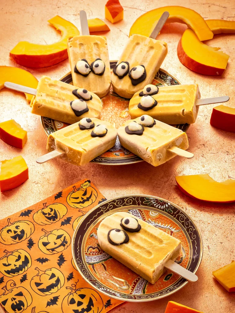 On a bronze-colored background, a small patterned plate with a Halloween popsicle with pumpkin lies in the foreground. Next to it is a napkin printed with pumpkin motifs. Behind it is a larger plate with six more Halloween popsicles. Surrounded by slices of sliced Hokkaido pumpkin.