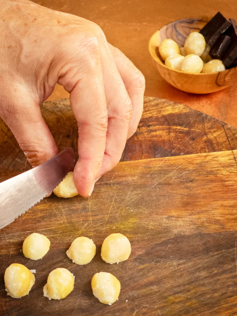 On a bronze-colored base is a wooden bowl with whole macadamia nuts and pieces of chocolate in the background. In front of it is a round wooden chopping board. One hand is holding a macadamia nut between the thumb an fingers, and a serrated knife is going to cut it in half. In the foreground are six macadamia nuts, already cut in half.