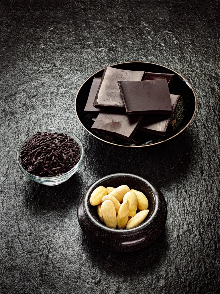 On a black slate background, three bowls with chocolate pieces, chocolate sprinkles and blanched almonds.