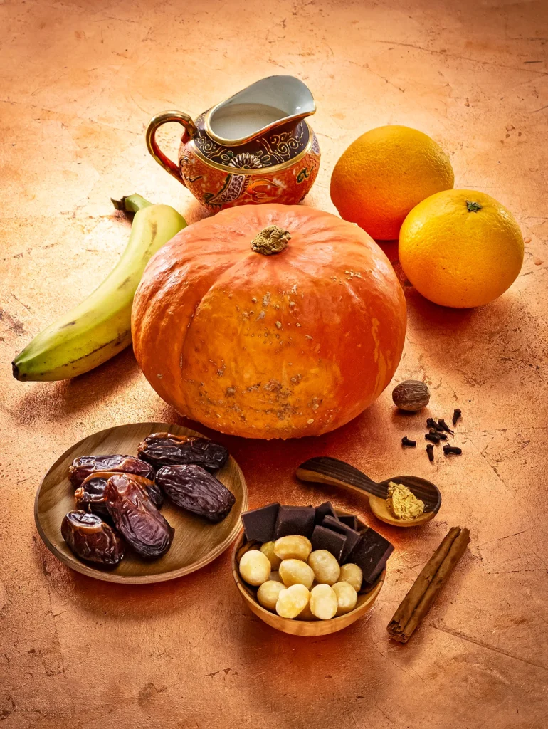 A whole Hokkaido pumpkin sits in the center of a bronze-colored base. In front of it is a small wooden plate with Medjool dates, and a wooden bowl with whole macadamia nuts and pieces of chocolate. There is also a wooden spoon with ginger powder, a cinnamon stick, nutmeg, and some dried cloves. Behind the pumpkin is a banana, a jug of coconut milk, and two oranges.