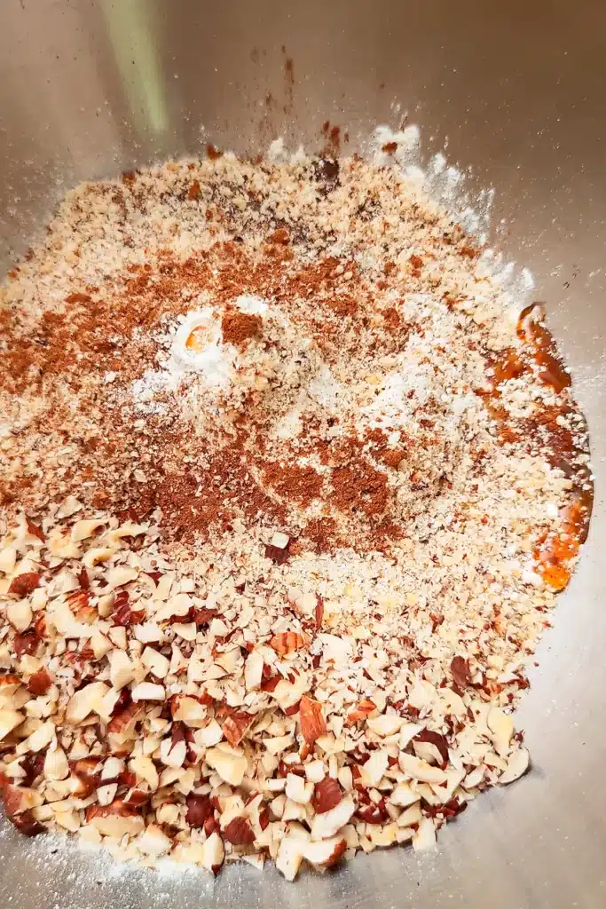 In a large stainless steel bowl is the mixture for the crumble dough - with maple syrup, ground hazelnuts, chopped hazelnuts, ceylon cinnamon.