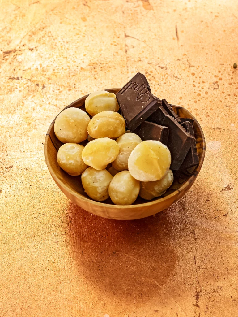 On a bronze-colored base is a wooden bowl with whole macadamia nuts and pieces of chocolate.