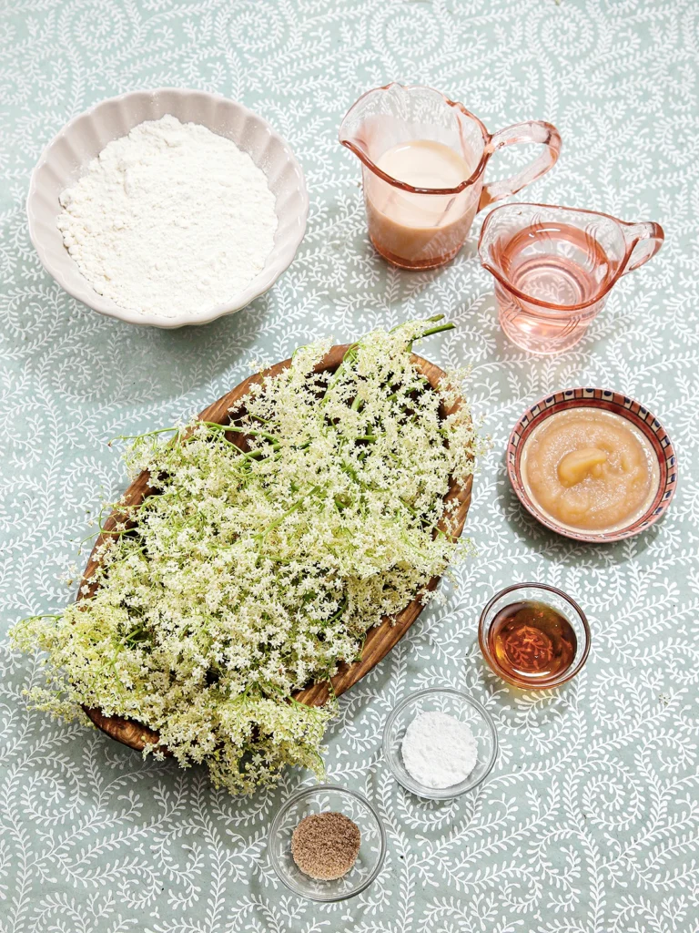 There are many elderflowers in a large wooden bowl. Around it are some bowls with spelled flour, applesauce, maple syrup, baking powder, and vanilla sugar. Next to the flour bowl there are two jugs with oat milk and soda. Everything is on a gray background with a white leaf pattern.