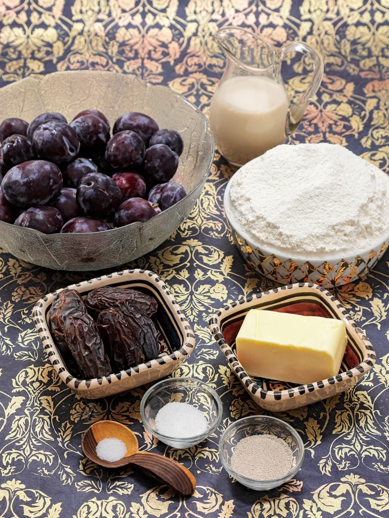 Bowls with fresh plums, spelt flour, dates, butter substitute, dry yeast and vanilla sugar stand on a patterned surface. Before that, a wooden spoon with a little salt. In the background a jug with oat milk.