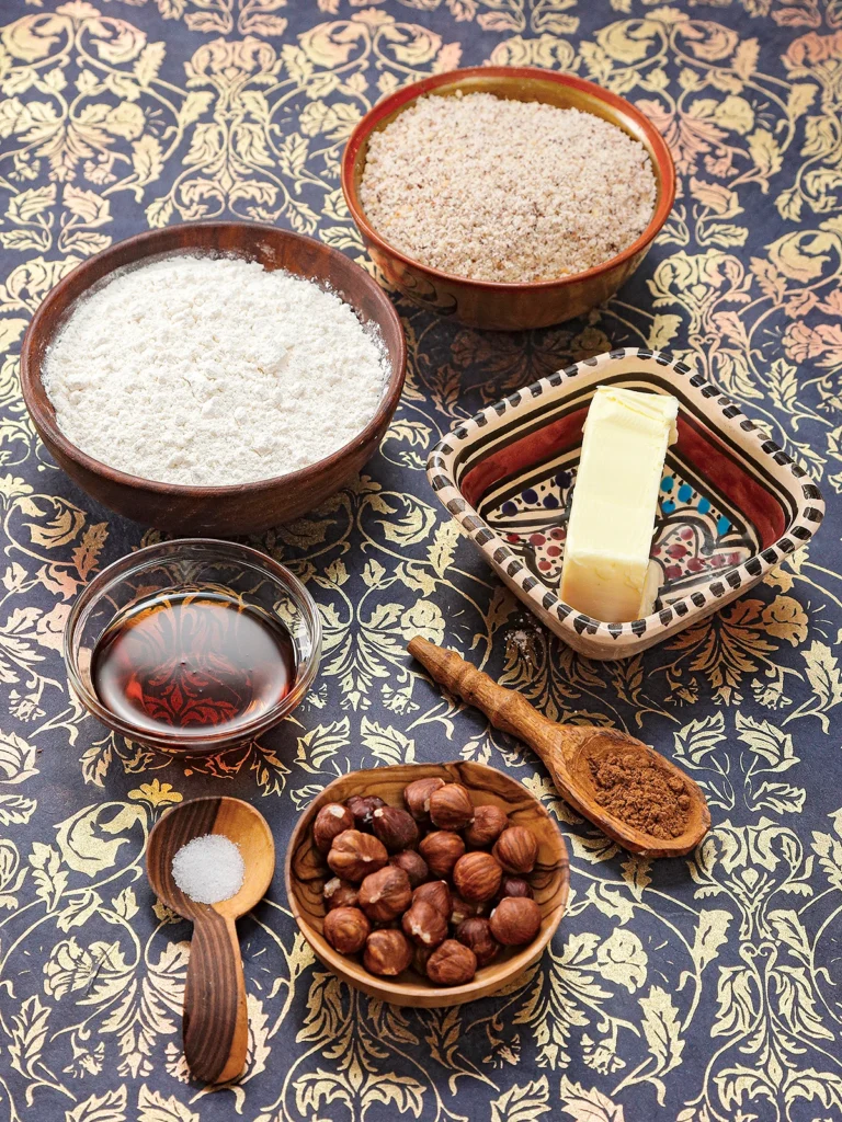 Bowls with flour, ground hazelnuts, butter substitute, maple syrup and whole hazelnuts stand on a patterned surface. In front of it are two different wooden spoons filled with Ceylon cinnamon and salt.