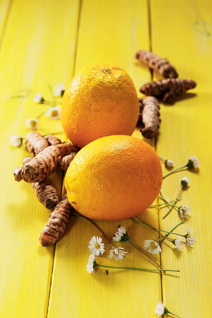 Two oranges lie on a yellow wooden background. Tumeric roots and daisies are spread around them.