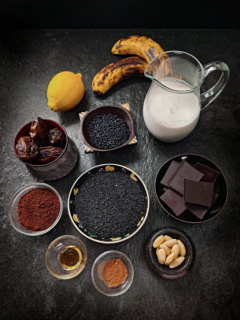 There are many bowls on a black slate background. In the middle, there is a bowl with black sesame, around that there are bowls with chocolate pieces, blanched almonds, cinnamon, sesame oil, cocoa powder, dates, Beluga lentils, and a jug with coconut milk. Behind it there a two ripe bananas and a lemon.