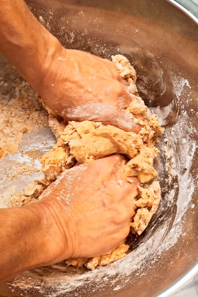 Two hands in a large stainless steel bowl begin to knead the ingredients for the crumble dough together.