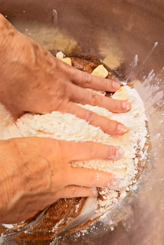 Two hands in a large stainless steel bowl begin to knead the ingredients for the yeast dough together.