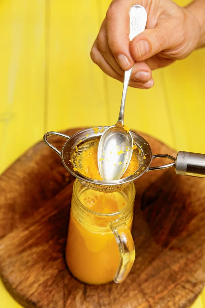 A jug of turmeric juice stands on a round wooden cutting board. A hand holds a spoon that squeezes the grated turmeric root into a small sieve that is placed over the jug. A yellow wooden background can be seen in the background.