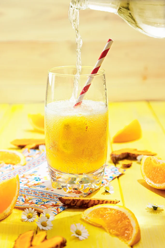 A bottle of sparkling water pours water in a glass with orange turmeric ice cubes and a straw stands on a yellow wooden base. Around the glass are slices of tumeric, oranges, daisies, and a patterned napkin.