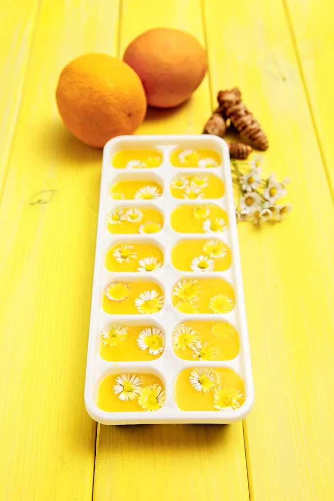 Prepared ice cube mold for orange turmeric ice cubes with daisy flowers stands on a yellow wooden underground. Behind the mold there are oranges, tumeric roots and some daisy flowers.