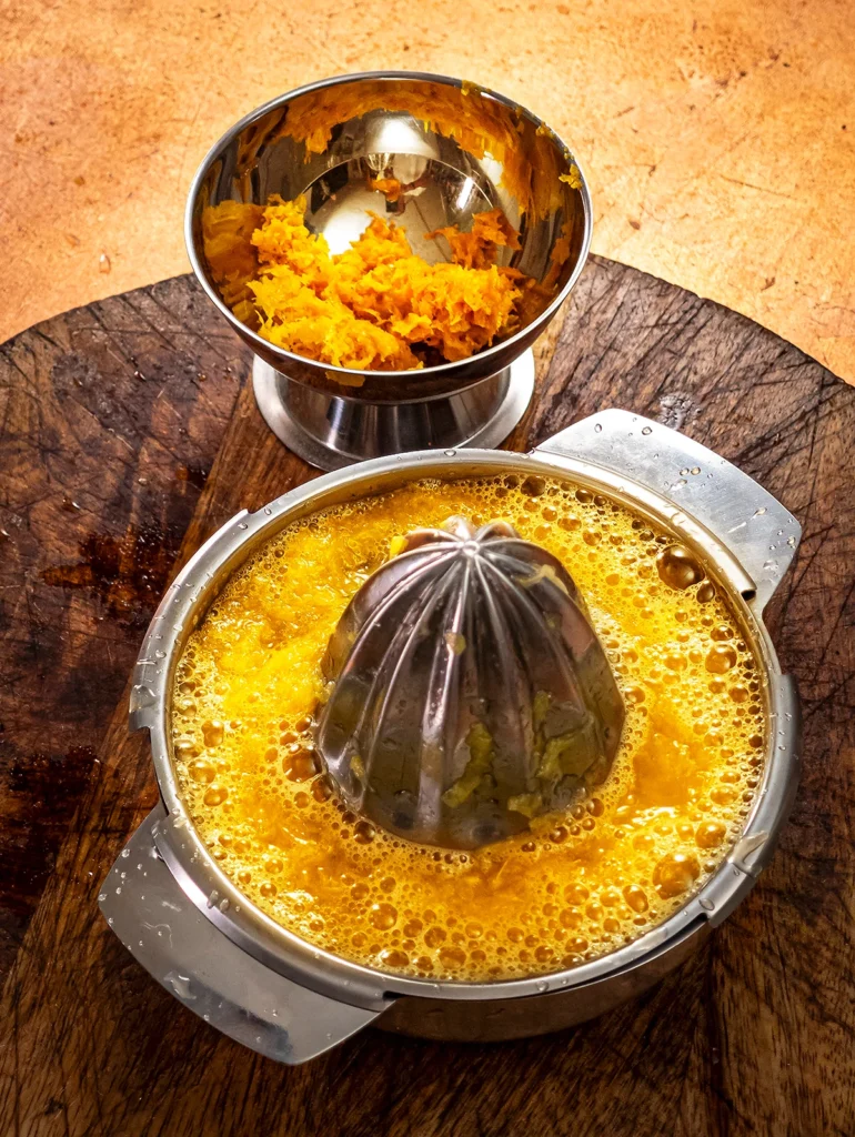 On a bronze-colored base stands a wooden cutting board with a stainless steel juicer filled with orange juice and a stainless steel bowl with grated orange peel.