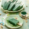 There is a protein popsicle on a green plate. On the left is a patterned napkin. On the other side is a small yellow-flowered plate with a small glass bowl filled with green spirulina powder. Behind it is a green serving plate with a lots of protein popsicles. Cashew nuts are scattered on the light green wooden base.