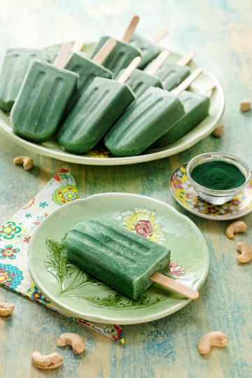 There is a protein popsicle on a green plate. On the left is a patterned napkin. On the other side is a small yellow-flowered plate with a small glass bowl filled with green spirulina powder. Behind it is a green serving plate with a lots of protein popsicles. Cashew nuts are scattered on the light green wooden base.