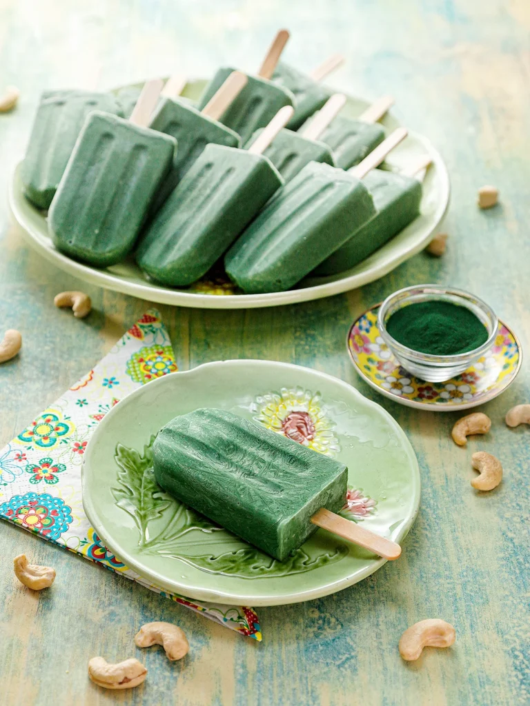 There is a protein popsicle on a green plate. On the left is a patterned napkin. On the other side is a small yellow-flowered plate with a small glass bowl filled with green spirulina powder. Behind it is a green serving plate with a lots of protein popsicles. Cashew nuts are scattered on the light green wooden base.