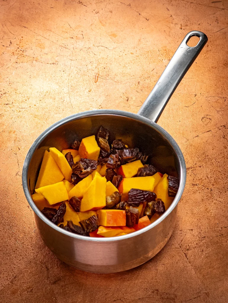 A stainless steel pot with pieces of Hokkaido pumpkin and dates stands on a bronze-colored base.