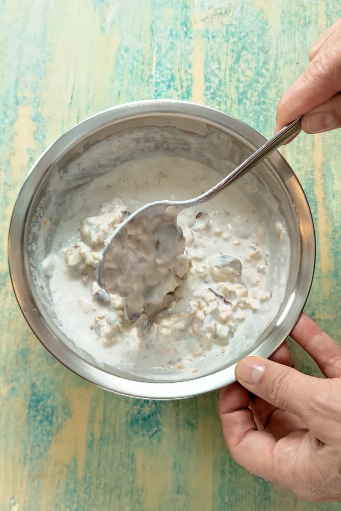 One hand holds a spoon and stirs in a stainless steel bowl, which is held by the other hand. The bowl contains a mixture of coconut milk, pieces of dates, and oat flakes for our protein popsicles. The bowl stands on a light green wooden underground.