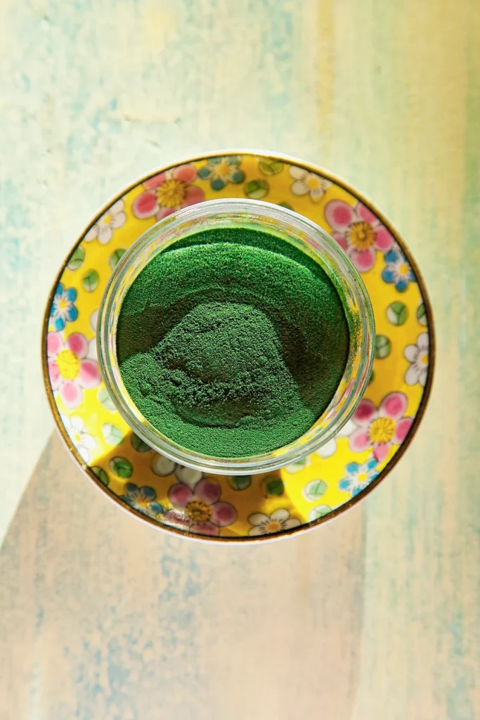 Top view of a small bowl of spirulina powder. The glass bowl is placed on a yellow flowered plate.