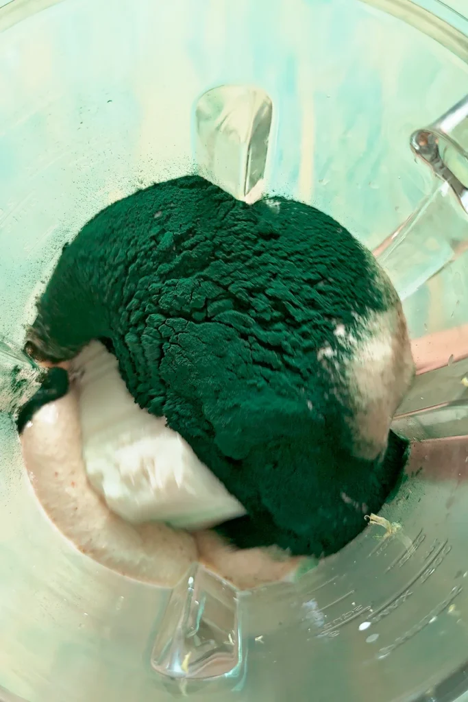 Top view of the glass container of a blender with all the ingredients for protein popsicles, with green spirulina powder on top.