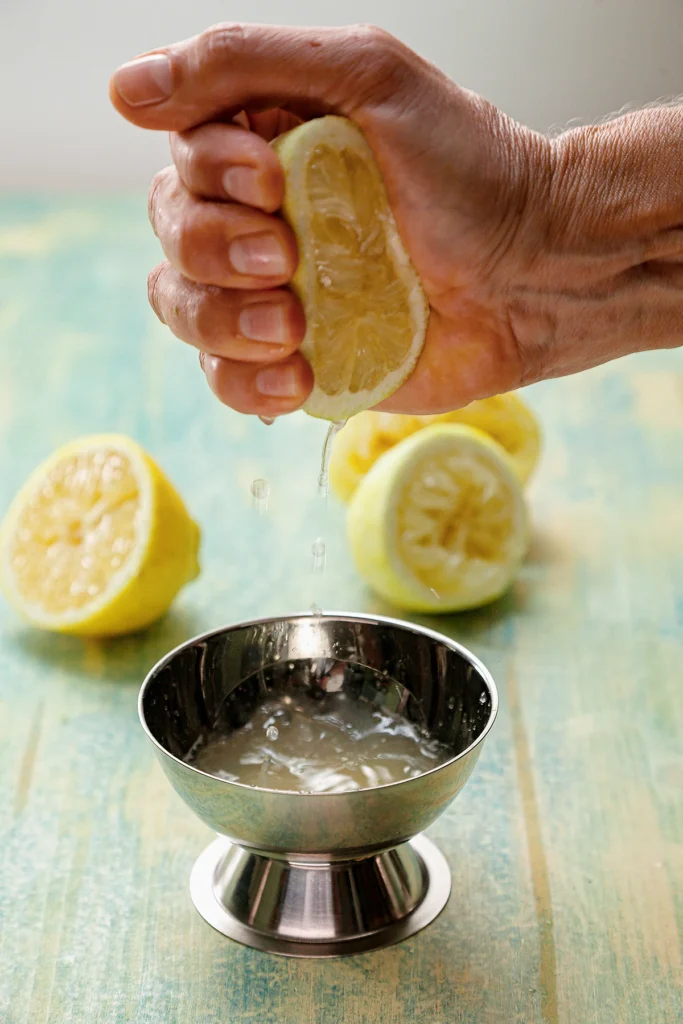 Half a lemon is squeezed with one hand. The juice is collected in a stainless steel bowl placed on a light green wooden base. In the background are three half-lemons.