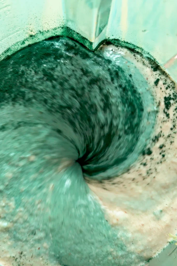 A top view of the glass container of a blender in which all of the ingredients for protein popsicles and the green spirulina powder have begun to be mixed.