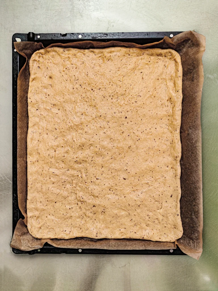 The yeast dough is completely spread out on the baking paper on size of the oven baking tray.