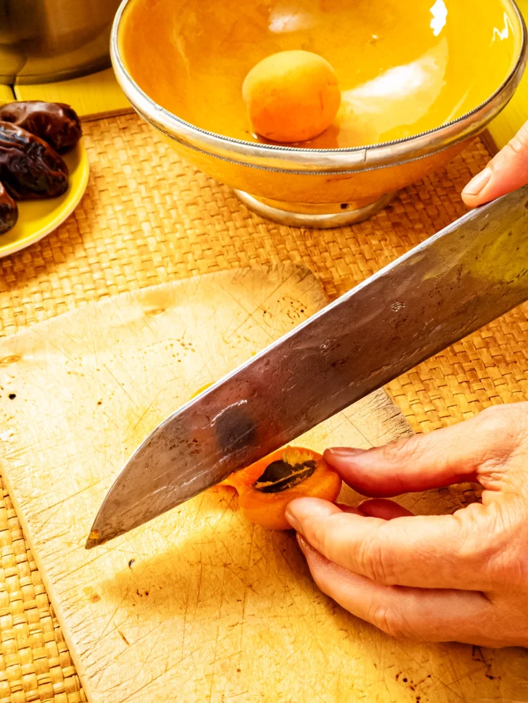 Two Hands hold a huge knife, and an apricot. The knife cut the apricot. The apricot lies on a wooden cutting board, that lies on a bast mat. In the background is a yellow bowl with an apricot, besides is a yellow plate with dried dates.