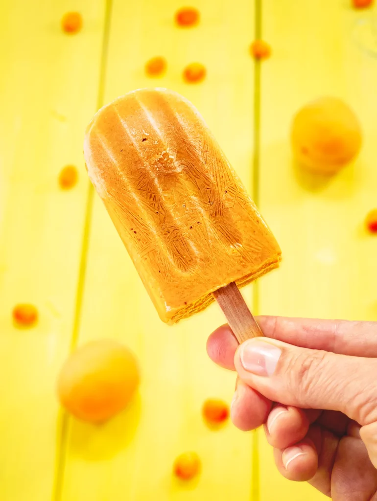 A hand holds a sea buckthorn popsicle. In the yellow backgroud there are blurred apricots and sea buckthorn berries.