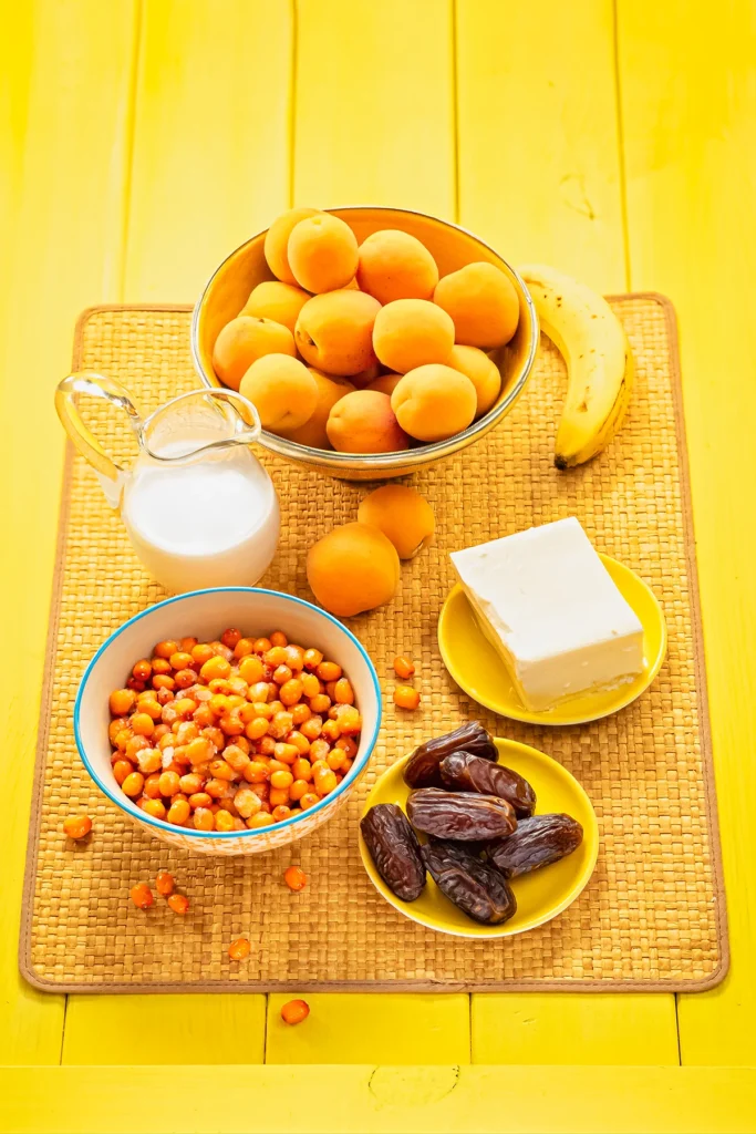 On a yellow colored wooden base there is a straw mat. On the mat are some bowls with thawed Sea buckthorn berries, fresh apricots and dried dates. Behind the bowls lies a ripe banana, a jug with coconut milk and a yellow plate with a block of silken tofu.