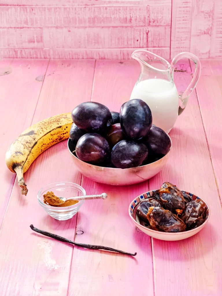 On a pink-colored base ther are some bowls with fresh plums, dried dates and cinnamon powder. In the foreground is a whole vanilla pod. Behind the bowls lies a ripe banana and a jug with coconut milk.