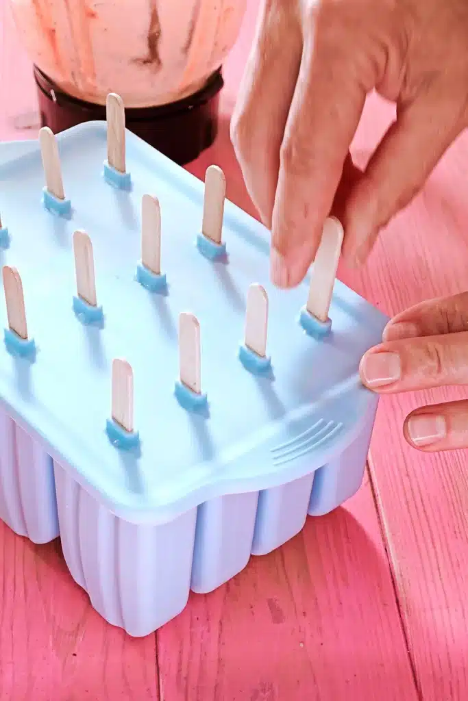 The silikon popsicle mold is closed with a lid and wooden sticks are inserted by hand. The blue mold stands on a pink wooden underground.