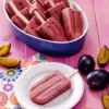 A small patterned plate with a plum popsicle stands in the foreground on a pink-colored base. Next to it is a napkin printed with floral motifs. Behind it is a large bowl with more plum popsicles. The plate and the bowl are surrounded by fresh plums, some are cut into slices.