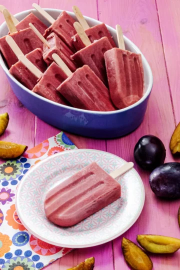 A small patterned plate with a plum popsicle stands in the foreground on a pink-colored base. Next to it is a napkin printed with floral motifs. Behind it is a large bowl with more plum popsicles. The plate and the bowl are surrounded by fresh plums, some are cut into slices.