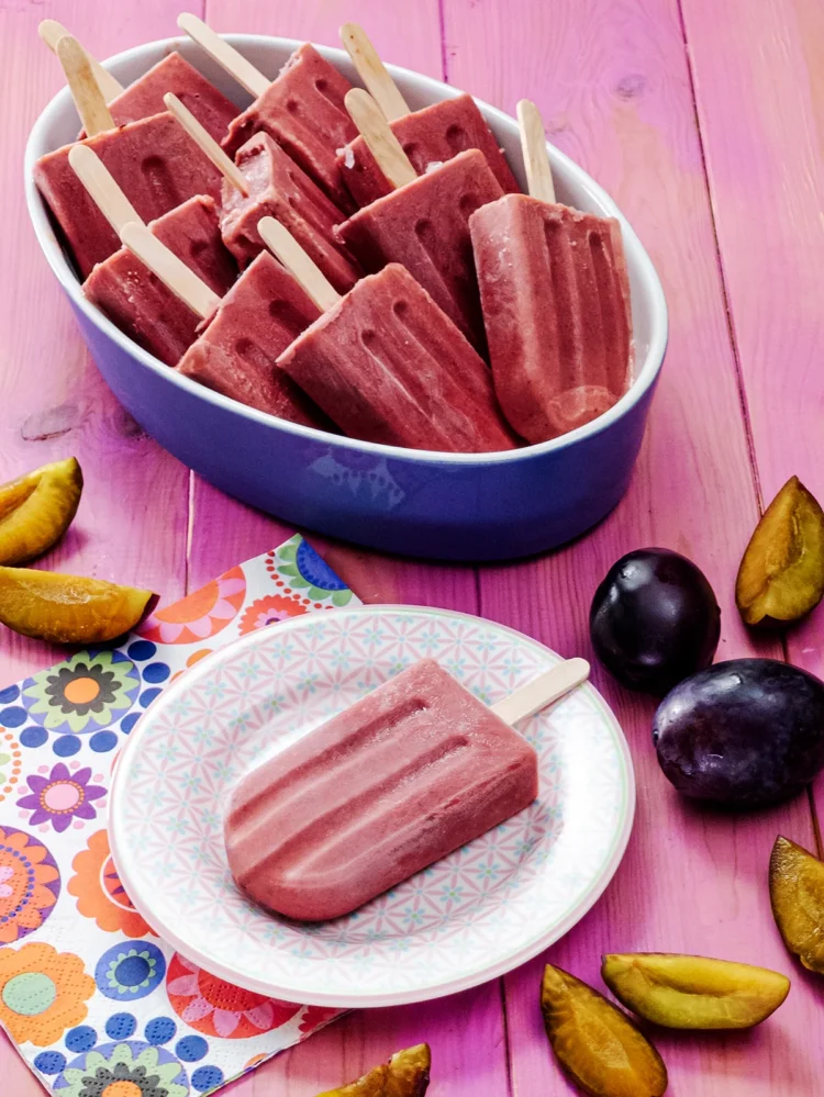 A small patterned plate with a plum popsicle stands in the foreground on a pink-colored base. Next to it is a napkin printed with floral motifs. Behind it is a large bowl with more plum popsicles. The plate and the bowl are surrounded by fresh plums, some are cut into slices.