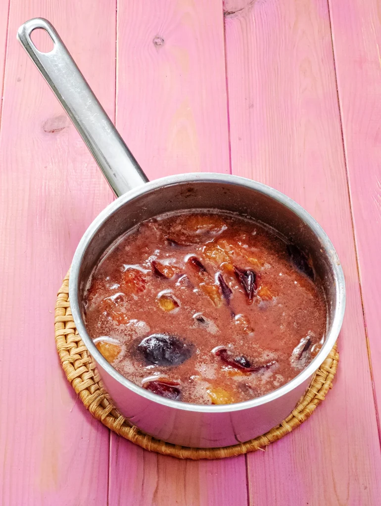 A stainless steel pot stands on a round seaweed saucer, the saucer with the pot stands on a pink base. The pot is filled with the cooked plum mixture.