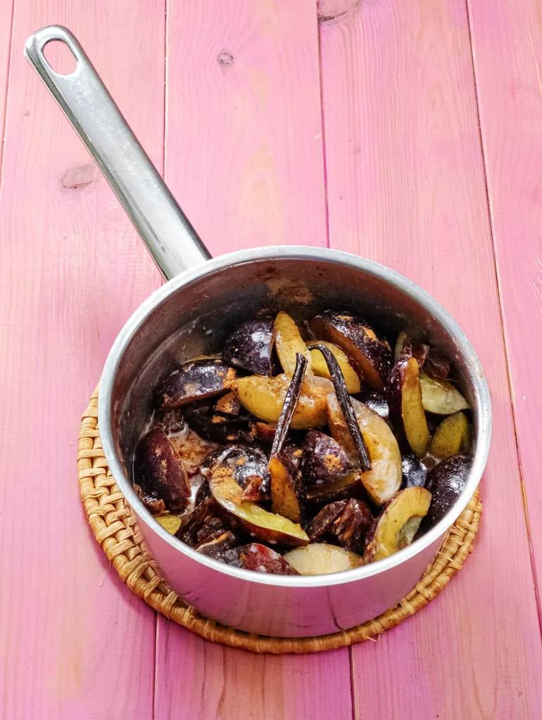 A stainless steel pot stands on a round seaweed saucer, the saucer with the pot stands on a pink base. The pot is filled with quartered fresh plums, coconut milk, cinnamon powder and two halves of a vanilla pod.