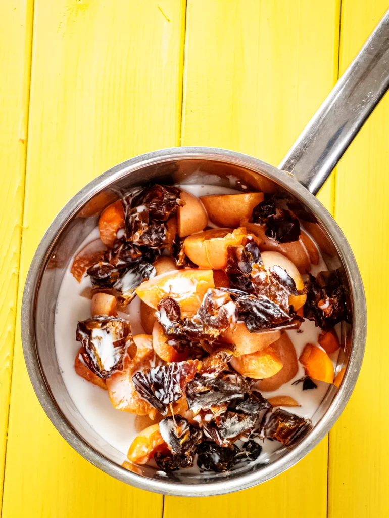 On a yellow wooden base stands a small stainless steel pot with a mixture of freshly cut apricots, dates and coconut milk.