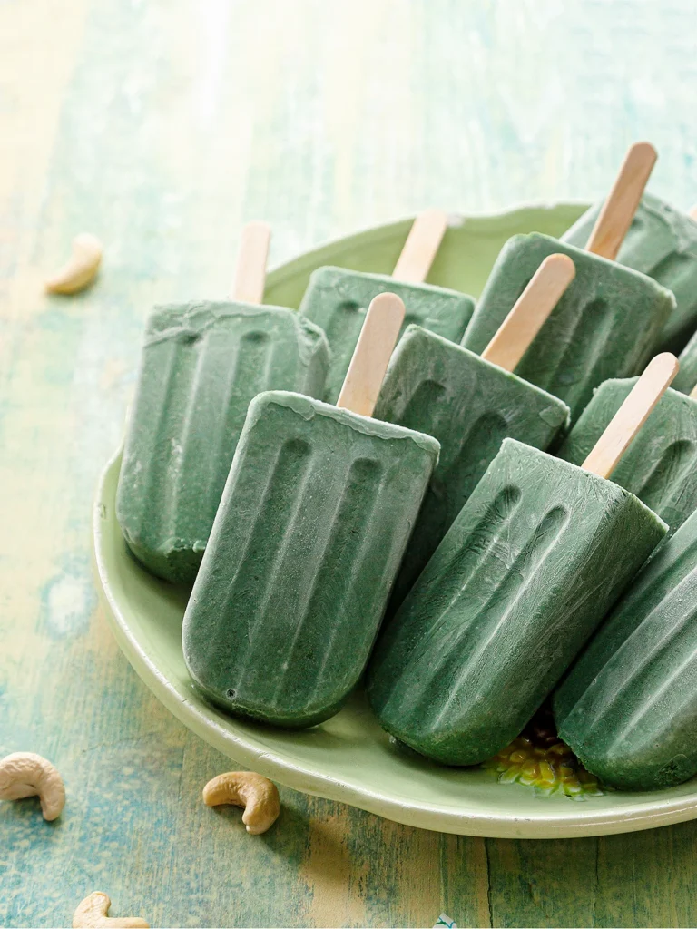 A green serving plate with lots of protein popsicles stands on a green wooden base with some cashew nuts around it.