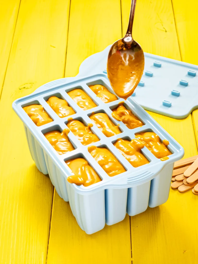 Filling the popsicles mold with the ready mixed sea buckthorn fruit mixture, with the help of a silver spoon. All compartments are full. Behind the mold is the lid. The mold stands on a yellow wooden base.