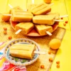 There is a sea buckthorn popsicle on a small colourful plate. On the left is a patterned napkin. Behind the small plate is an orange serving plate with a lots of sea buckthorn popsicles. The plates are placed on a grass mat. Apricots and sea buckthorn berries are scattered on the yellow wooden base and the mat.