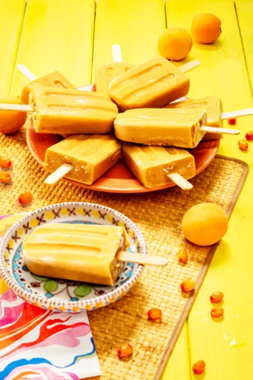 There is a sea buckthorn popsicle on a small colourful plate. On the left is a patterned napkin. Behind the small plate is an orange serving plate with a lots of sea buckthorn popsicles. The plates are placed on a grass mat. Apricots and sea buckthorn berries are scattered on the yellow wooden base and the mat.